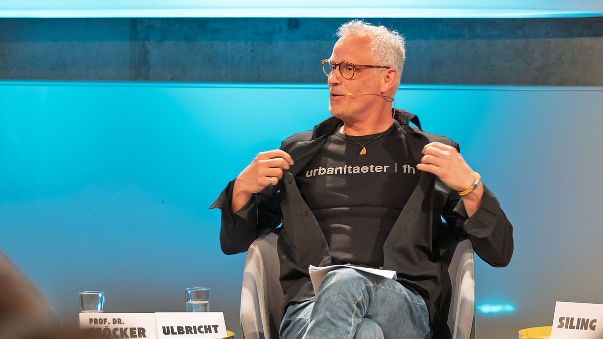 Männliche Person im Podium öffnet ihre Jacke und zeigt ein Shirt mit der Aufschrift "Urbanitaeter".