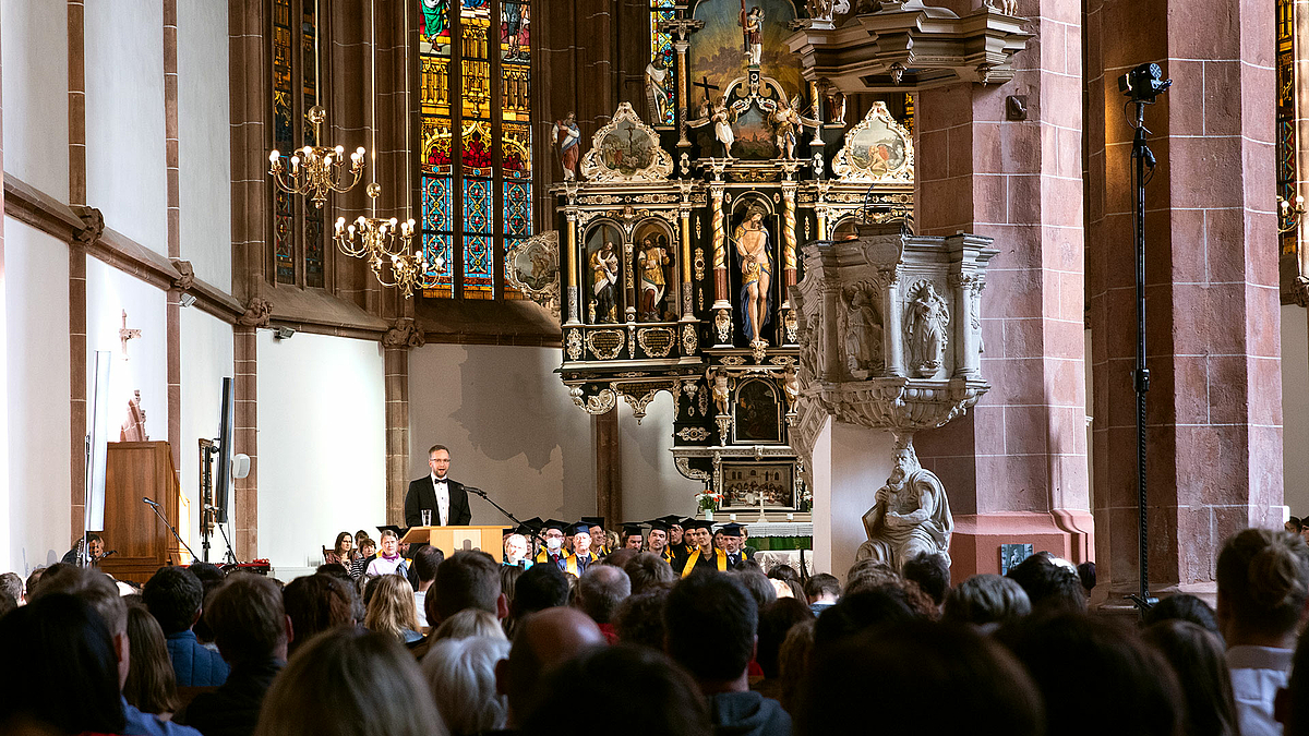 Studierende und Angehörige sitzen auf den Kirchenbänken der Mittweidaer Stadtkirche. Im Hintergrund glitzert der imposante Altar golden.
