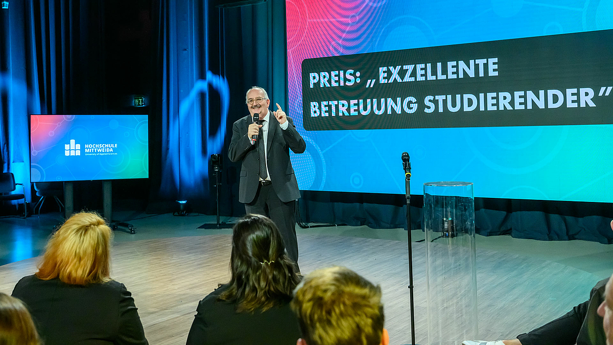 Eine mönnliche Person steht in der Studiokulisse vor Publikum, das von hinten zu sehen ist. "Preis für exzellente Betreuung Studierender" ist auf einem großen LED-Screen im Hintergrund zu lesen.