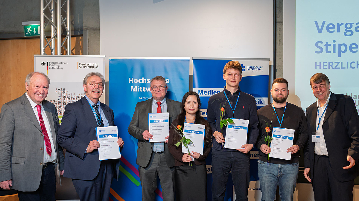 Fünf Personen mit Urkunden zwischen den beiden Übergebenden.