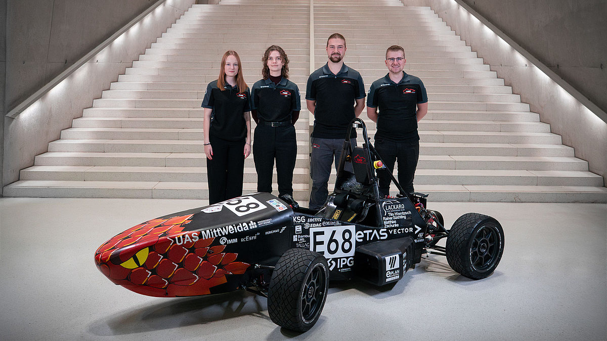Zwe i junge weibliche und zwie junge männliche Personen in TMM-Polos vor einer großen Treppe, vor ihnen steht das Rennauto.