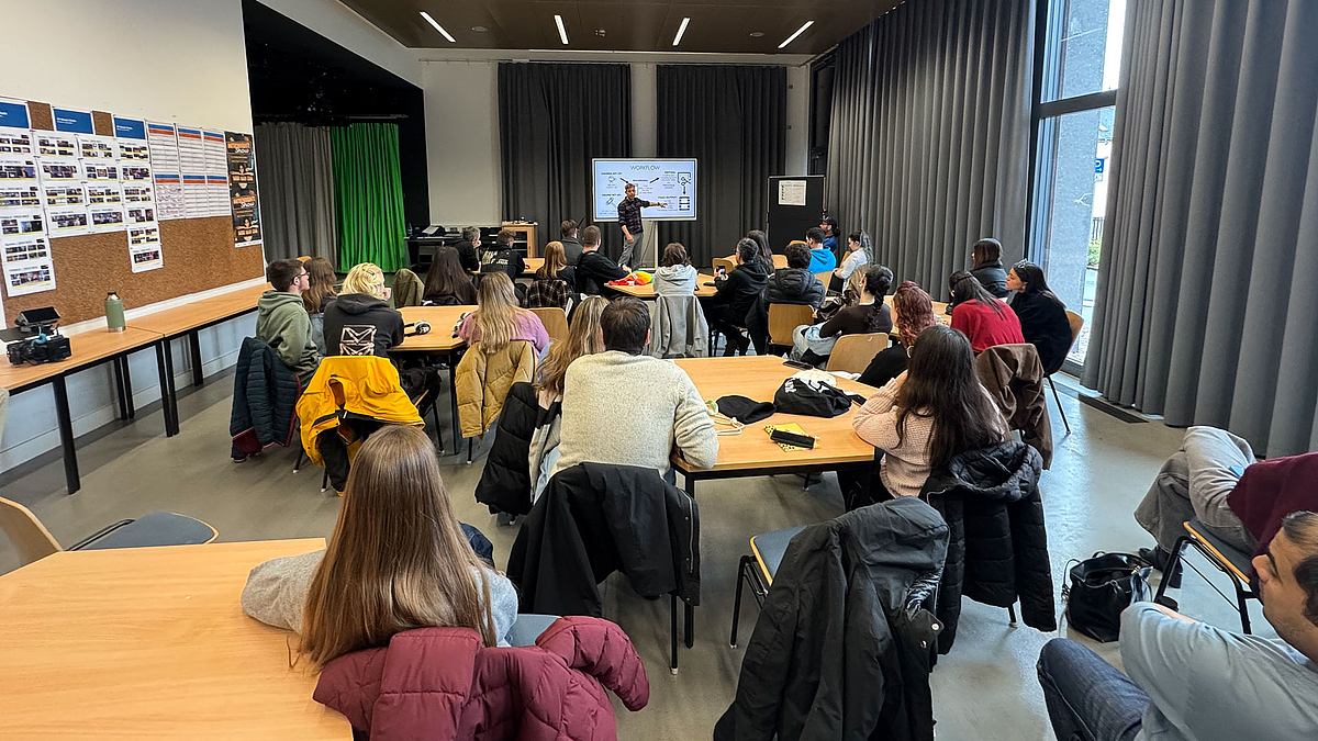 Uahlreiche junge Menshcn in einem großen Seminarraum schaen auf einen Dozenten vor einem großem Monitor.