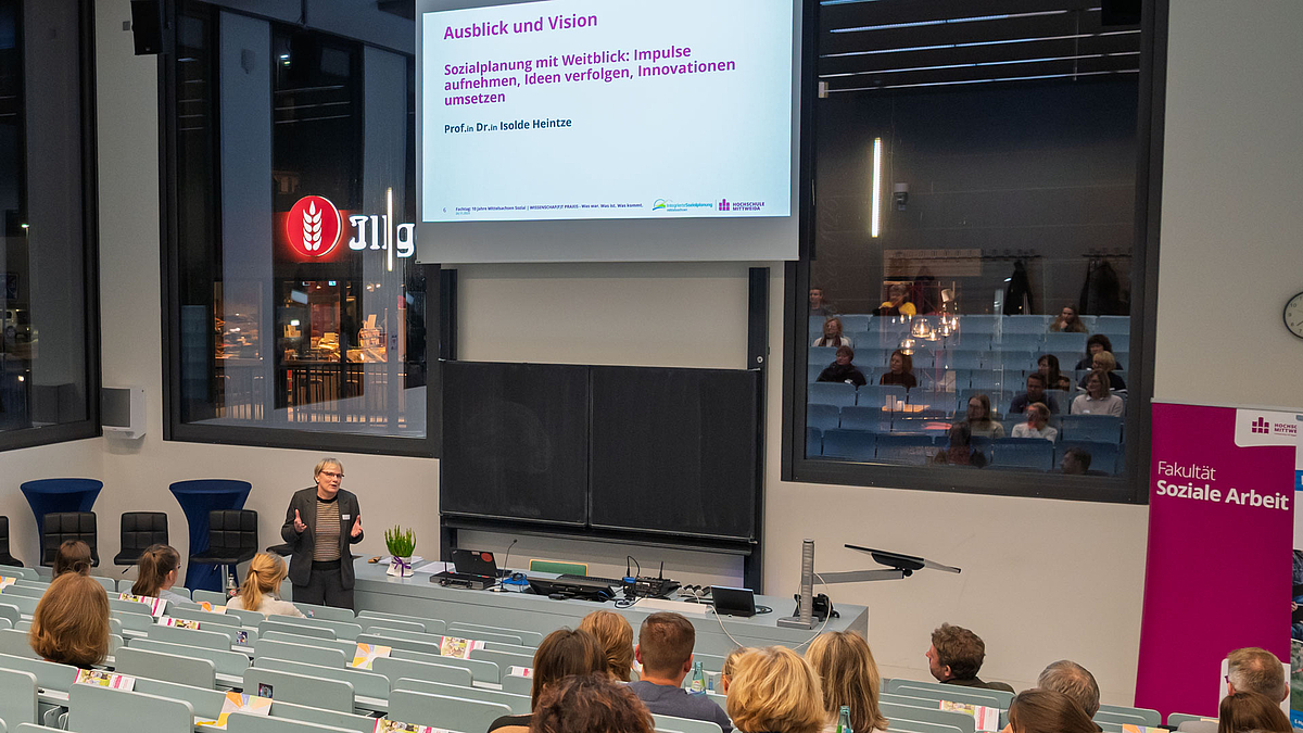 Weibliche Person spricht zum Publikum im Hördaal. Auf der Leiwnand über ihr ist zu lesen: "Ausblick und Vision Sozialplanung mit Weitblick: Impulse aufnehmen, Ideen verfolgen, Innovationen umsetzen."