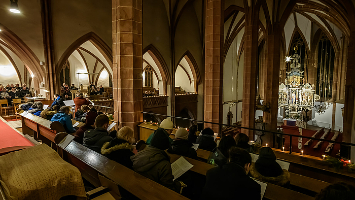 Blick in das spätgotische Kirchenschiff. Im Hintergrund der erleuctete Altarrum, im Vordergrund Menschen auf Kirchenbänke auf ein Emporen der Kirche.