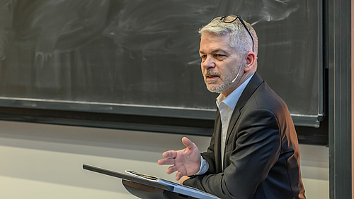Prominent und kompetent: Dr. Carlo Masala, Professor für Internationale Politik an der Universität der Bundeswehr, bei der dritten von sieben Veranstaltungen der Öffentlichen Ringvorlesung „Zeiten wenden“ an der Hochschule Mittweida. Eine männliche Person mit entschiedener Mine vor einer schlecht geputzten Tafel, die Brille nach oben ins Haar geschoben, den linken Ellbogen auf ein Rednerpult abgestützt und die rechte Hand geöffnet, spricht zu einem im Bild nicht sichtbaren Publikum.