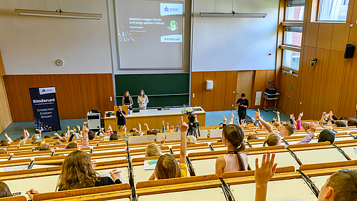 Robin Biebl vom Kinderuni-Team der Hochschule Mittweida (im Bild mit Mikrofon) begrüßt rund 150 Kinder zur Vorlesung im großen Hörsaal.