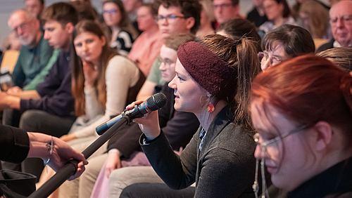 Viele Personen auf Stühlen. Eine junge weibliche Person spricht in ein Mikrofon, das ihr gereicht wird. 