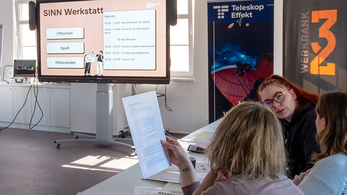 Workshop vor dem Gründungsforum (Foto: Olivia Weinehl) : Drei Personen im Gespräch. Im Hintergrund ist ein großer Screen zu sehen, auf dem „SINN Werkstatt“ zu lesen ist.