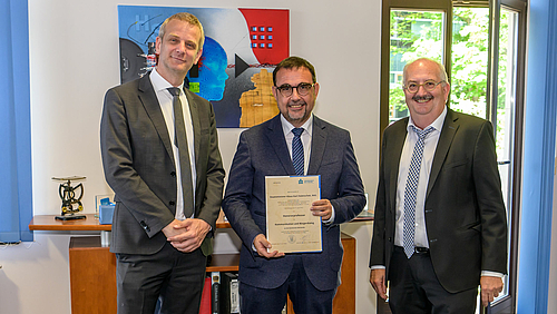 In die Mitte genommen: Professor Klaus Holetschek (Mitte) mit Medien-Dekan Markus Heinker (l.) und Rektor Ludwig Hilmer. Das Foto zeigt drei männliche Personen. Die Person in der Mitte hat eine Urkund eine Urkunde in der Hand.