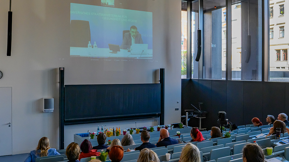 Blick von hinter über die Köpfe der Menschen im Hörsaal nach vorn. An die Rückwand ist ein Video mit einer an einem Pult sitzenden männlichen Person projiziert, die vor einem Laptop sitzt.