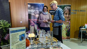 Zwei weibliche Personen stehend an einem Tisch, auf dem verschiedene Gefäße stehen, Im Hintergrund stehen zwei Rollups mit Naturmotiven.