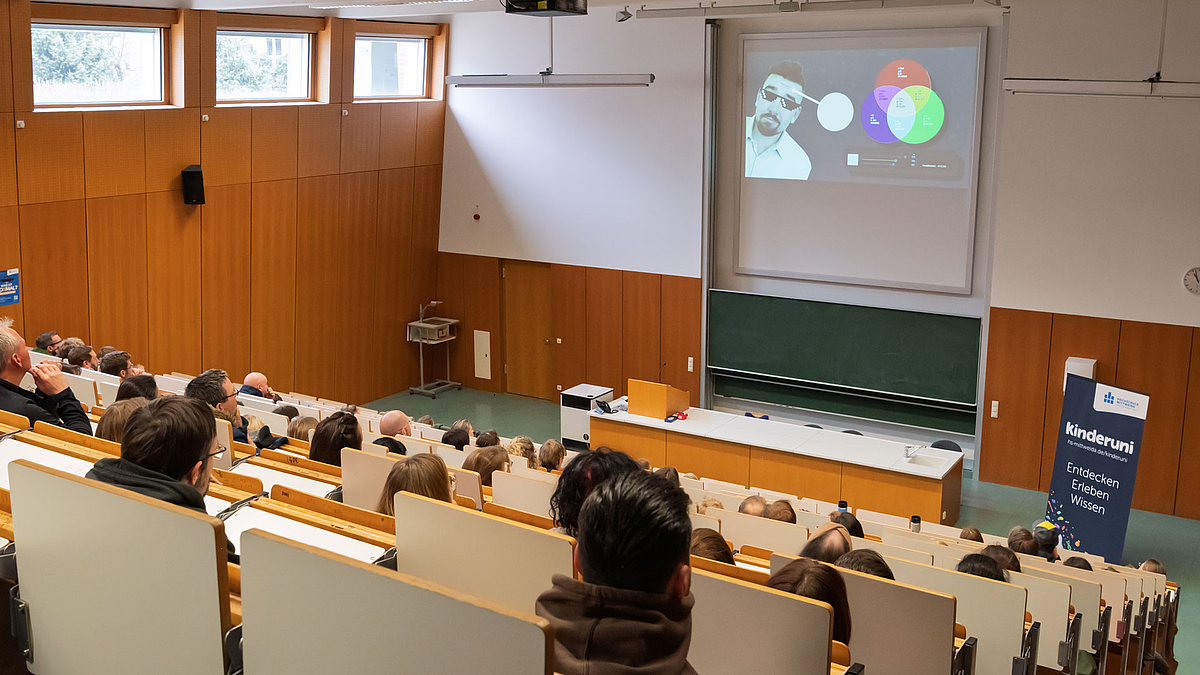 Blick auf das Beamerbild im Elternhörsaal, auf dem Dozent mit Sonnenrille neben einem RGB-Farbmodell zu sehen ist.
