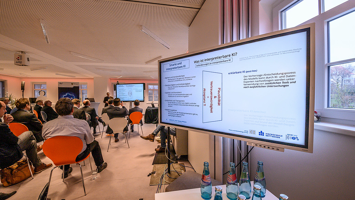 Das Foto zeigt rechts einen großen Monitor mit einer Powerpoint-Folie zu smarter KI. Links im Bild sind Teilnehmende von hinten auf Stühlen zu sehen. Im Bildhintergrund am Raumende der Referent neben einem weiteren Display mit derselben Folie.