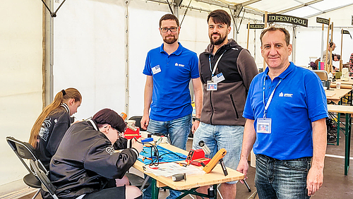 Professor Michael Kuhl, sein Mitarbeiter Markus Süß und Masterstudent Steve Kriebel (v.r.n.l.) am Stand der Hochschule Mittweida bei der „beam! – be a maker“. In einem Messezelt stehen zahlreiche Tische und Bänke. Drei männliche Personen schauen in die Kamera. Zwei von ihnen tragen blaue „Hochschule Mittweida“-Polohemden. Vor ihnen an einem Tisch sitze¬n zwei Jugendliche vertieft in Arbeiten mit dem Lötkolben.