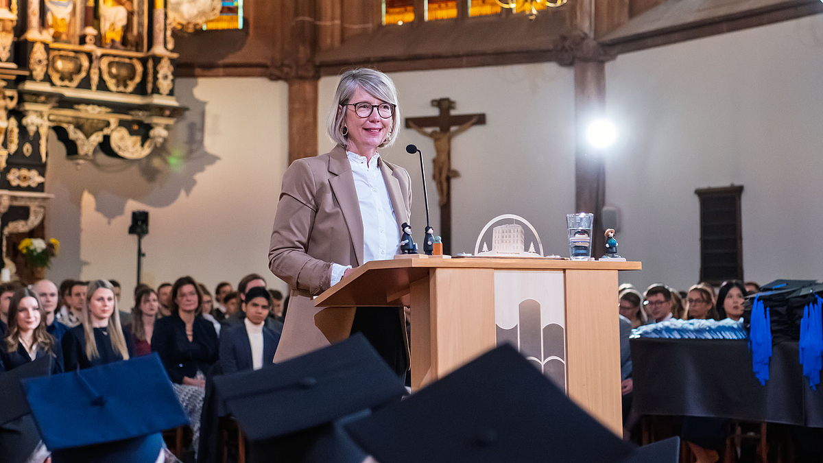 Selbe Person wie im Bild zuvor am Rednerpult miit Absolvent:innen hinter sich im Altarraum.