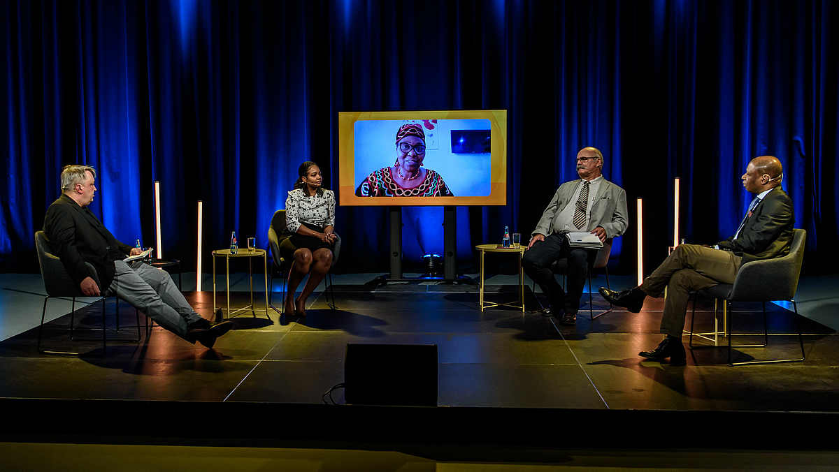 Das Foto zeigt das Podium mit dem Moderator links und den drei Teilnehmenden im TV-Studio: Nessma Adil Yousif, Prof. Dr.  Andreas Wrobel-Leipold und Dr. Gabin Ananou. Die vierte Teilnehmerin, Dr. Hedwig Ngwa aus Kamerun, ist auf einem Monitor in der Mitte hinter dem Podium zu sehen.