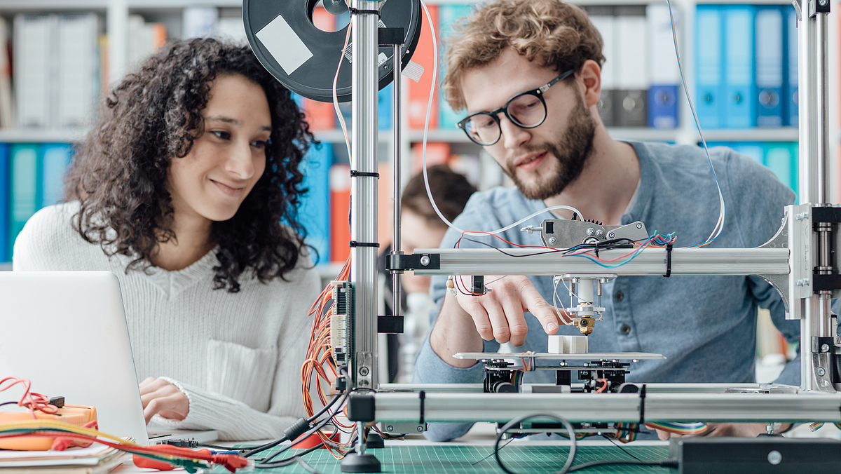 Eine Ferua und ein Mann sitzen lächelnd vor einem 3D-Drucker, der Mann deutet mit dem Finger auf den Drucker.
