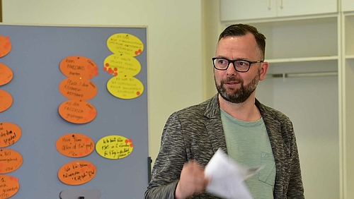 Eine männliche Person steht referierend mit einem Blatt Papier in der Hand vor eine Pinnwand mit Moderationskarten und schaut in die linke Bildhälfte- Der Inhalt der Moderationskarten ist nicht zu erkennen.