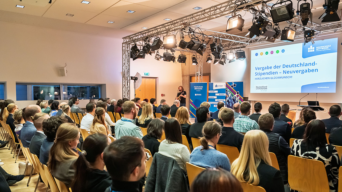 Derselbe Musiker mit dem Publikum im Vordergrund. Im Hintergrund die drei blauen Rollups und die Leinwand auf die eine Folie mit dem Text „Vergabe der Deutschlandstipendien – Neuvergaben“ und den Logos von Hochschule und Deutschlandstipendium projiziert ist.