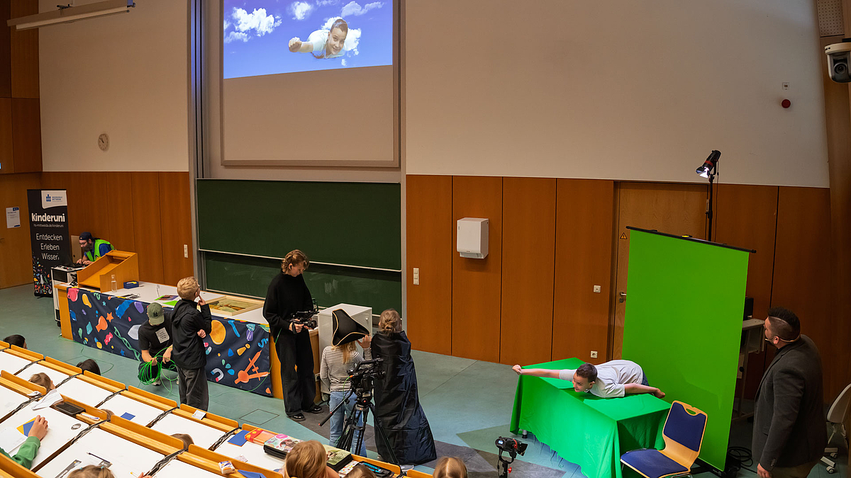 Ein Junge liegt mit ausgestrecktem Arm auf einem grünen Tisch vor einem grünen Hintergrund. Derselbe Junge ist auf dem Beamerbild vor einem blauen Himmel mit Wolken zu sehen.