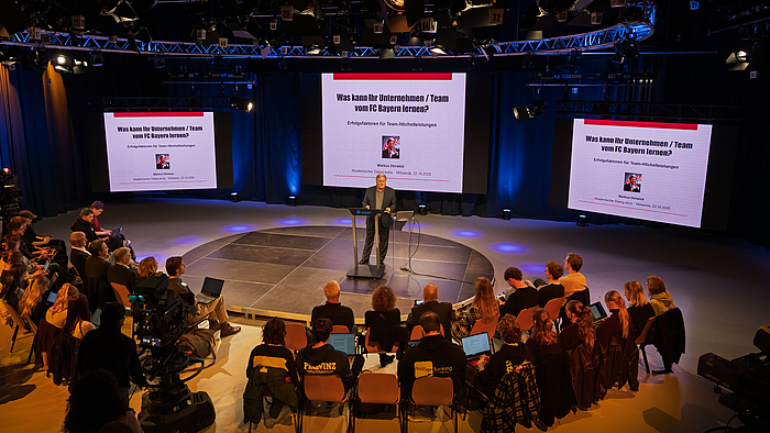 Großes Thema im großen Studio. Beim Akademischen Dialog Mittweida geben Branchenprofis ihr Wissen weiter. Blick von oben über die Köpfe hinweg ins TV-Studio. In der Mitte der Studiofläche spricht eine männliche Person an einem Rednerpult stehend. Hinter ihm auf drei großen LED-Screens ist das Thema des Vortrags zu lesen: "Was kann Ihr Unternehmen/Team vom FC Bayern lernen. Erfolgsfaktoren für Team-Höchstleistungen".