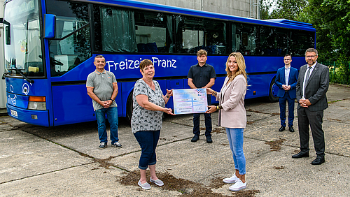 Das Foto zeigt vier männliche und zwei weibliche erwachsene Personen untschiedlichen Alters vor einem blauen Bus mit der Aufschrift "Freizeit Franz". In der Bildmitte vorne halten die zwei Frauen einen großen symbolischen Spendenscheck.