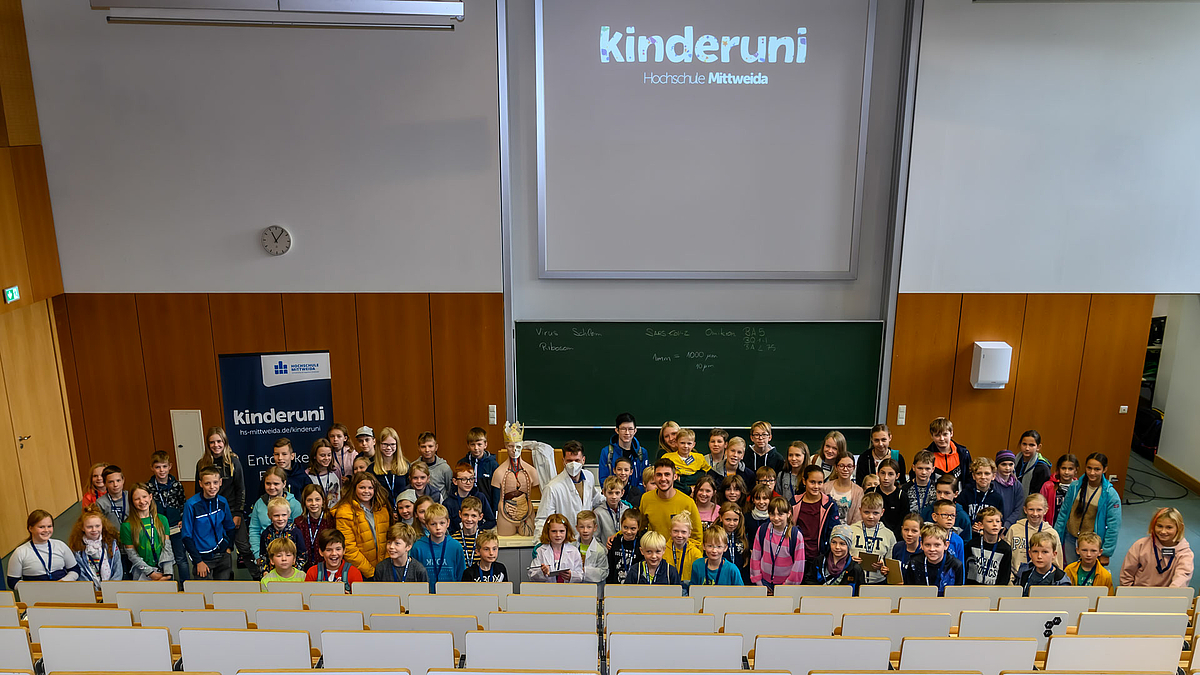 Alle Kinder der Vorlesung sowie Erwachsene des Kinderuni-Teams stehen dichtgedrängt am unteren Ende des Hörsaals. Sie füllen fast die gesamte Breite des Hörsaals und schauen nach oben zum Betrachtenden.