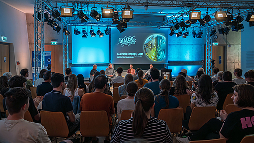 In einem großen Raum fällt der Blick über die Köpfe der Zuschauer:innen hinweg auf das mit vier Pesonen bestzte Podium unter an Traversen hängenden Scheinwerferen. Auf der Leinwand hinter ihnen ist das "Dialog Kontrovers"-Motiv und das Thema "Das ewige hybride Leben" projiziert.