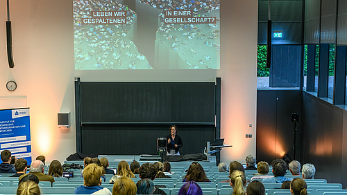 In der 5. Vorlesung innerhalb der Reihe „Zeiten wenden! Aussitzen – Analysieren – Anpacken“ an der Hochschule Mittweida gab Clara Dilger vom Forschungsinstitut Gesellschaftlicher Zusammenhalt (FGZ) tiefe Einblicke in aktuelle Forschungsergebnisse. Blick über die Köpfe der Zuhörenden im Hörsaal hinweg auf die Dozentin vorne am Pult. Über ihr ist eine Folie projiziert, auf der vor dem Hintergrund einer tiefen Erdspalte der Text „Leben wir in einer gespaltenen Gesellschaft“ steht.