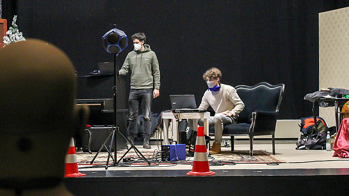 Das Foto zeigt zwei junge Männer mit FFP2-Masken und Laptops auf der Bühne des Theaters. Die Kulisse ist spärlich: ein Klavier, ein Sofa, ein Tisch sind zu sehen. Beherrscht wird das Motiv durch zwei orange-weiße Verkehrsleitkegel am Bühnenrand sowie einem Dodekaeder-Lautsprecher auf einem Stativ und Kabel. Im Bildvordergrund befindet sich im Anschnitt und leicht unscharf ein Kunstkopf.