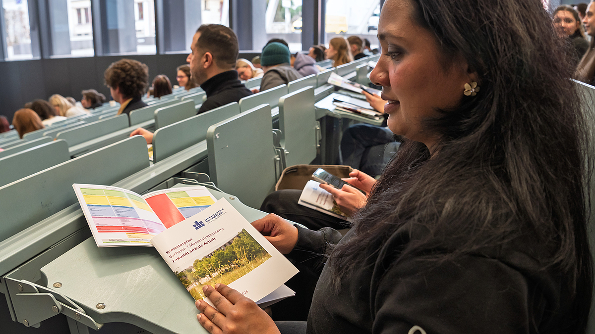 Eine weibliche Person in einer Hörsaalbank hat zwei Broschüren vor sich, eine trägt den Titel: „Semesterplan Bachelor-/Masterstudiengang. Fakultät Soziale Arbeit.“