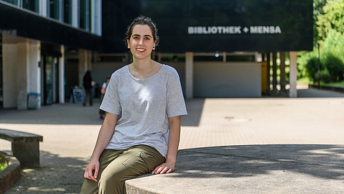 Abschied aus Mittweida: Ane Zuniga Gurruchaga verlässt die Hochschule nach zwei Semestern im Doppelabschlussprogramm mit neuen Lehrinhalten, viel Praxiserfahrung, dem Bachelorabschluss und neuen Freunden. Das Fotos zeigt eine lächelnde junge Frau, die Studentin Ane Zuniga Gurruchaga, auf einem großen Tisch aus Beton sitzen. Im Hintergrund ist ein Gebäude zu erkennen mit dem Schriftzug "Bibliothek + Mensa".
