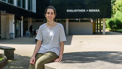 Abschied aus Mittweida: Ane Zuniga Gurruchaga verlässt die Hochschule nach zwei Semestern im Doppelabschlussprogramm mit neuen Lehrinhalten, viel Praxiserfahrung, dem Bachelorabschluss und neuen Freunden. Das Fotos zeigt eine lächelnde junge Frau, die Studentin Ane Zuniga Gurruchaga, auf einem großen Tisch aus Beton sitzen. Im Hintergrund ist ein Gebäude zu erkennen mit dem Schriftzug "Bibliothek + Mensa".