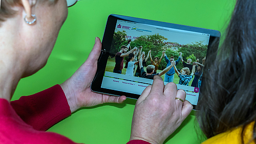 Neue aktuelle Einblicke auf die Lebenslagen der Menschen in Mittelsachsen im Blog „soziales.mittelsachsen.gemeinsam.machen“. Blick über die Schultern zweier Personen auf ein Tablet in den Händen der linken Person. Sie zeigt mit einem Finger der rechten Hand auf die Oberfläche des Tablets. Es zeigt eine Webseite mit einem Bildmotiv mit Menschen unterschiedlichen Geschlechts und Alters, die im Kreis stehend sich an den Händen fassen. Über das Bildmotiv ist zweimal der Schriftzug mit unterschiedlicher Deckkraft „soziales.mittelsachsen.gemeinsam.machen“ gelegt.