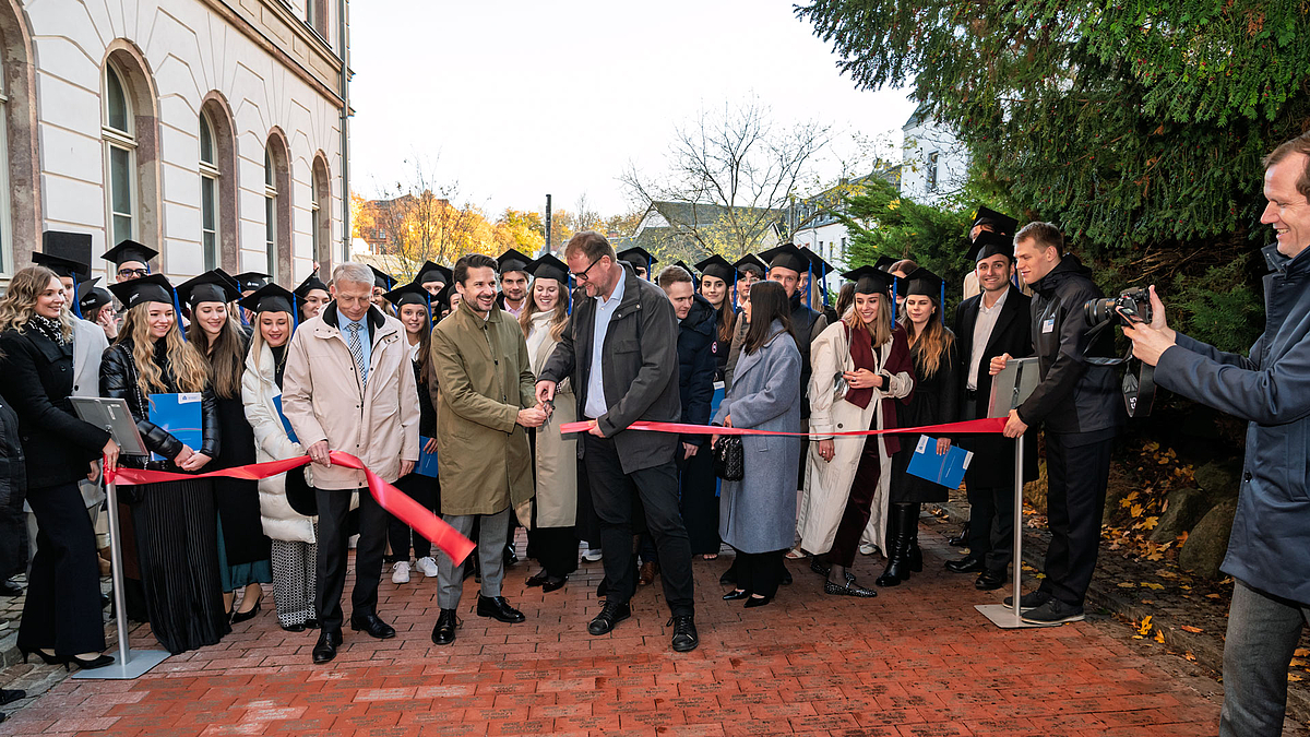 Drei männliche Personen auf einem gepflasterten Weg durchschneiden ein rotes Band. Hintter ihnen stehen zahlreiche Absolvent:innen.