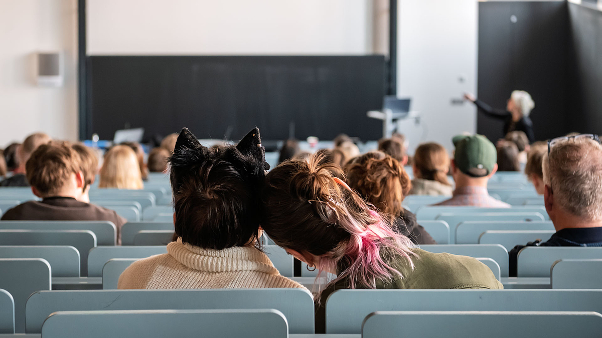 Blick von hinten über die Köpfe der Zuhörenden hinweg auf das untere Ende des Hörsaals. Im Fokus des Fotos sind zwei Personen in einer hinteren Bankreihe, deren Köpfe aneindandergeschmiegt sind. Eine der Personen trägt einen Haarreif mit Katzenohren. d