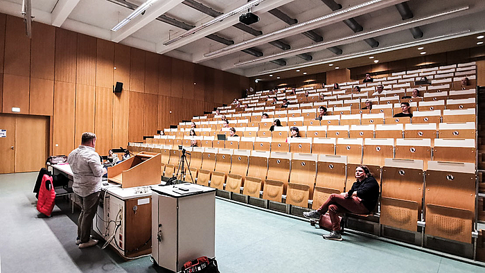 Das Fotos zeigt einen Hörsaal mit wenigen in großem Abstand voneinander sitzenden Studieren. Im Bildvordergrund ist ein Referent am Pult von hinten zu sehen. In der ersten Reihe sitzt rechts eine männliche Person. 