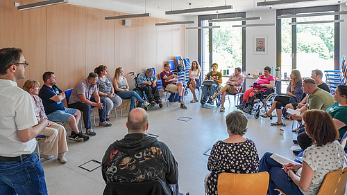 Im Studienmodul „Bewältigung von Lebenslagen“ bringt Professor Sebastian Noll (l.) Studierende und Klient:innen zusammen. (Foto: Christian Kästner) Eine Gruppe von rund 20 Menschen sitzt in einem Seminarraum in einem Kreis, die meisten auf Stühlen, zwei in einem Rollstuhl. Eine männliche Person steht am linken Bildrand der Gruppe zugewandt.