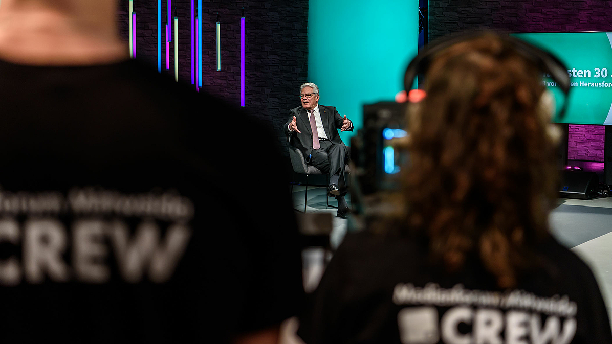 Das Foto zeigt Joachim Gauck im Hintergrund als Teilnehmenden des Podiums. Im Vordergrund sind unscharf zwei Studierende der Studio-Crew von hinten zu sehen.