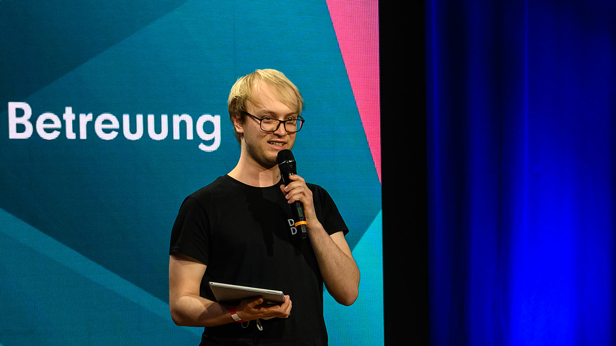 Das Foto zeigt einen jungen Mann mit Mikrofon. Auf dem Display im Hintergrund ist das Wort Betreuung zu lesen.