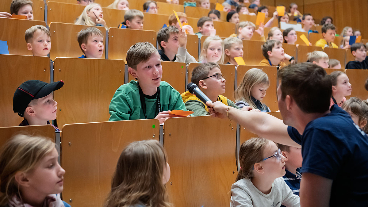 Ein Junge in einer Bankreihe unter vielen adneren Kindern. Er spricht in ein Mikrofon, das ihm der Moderator entgegenhält.