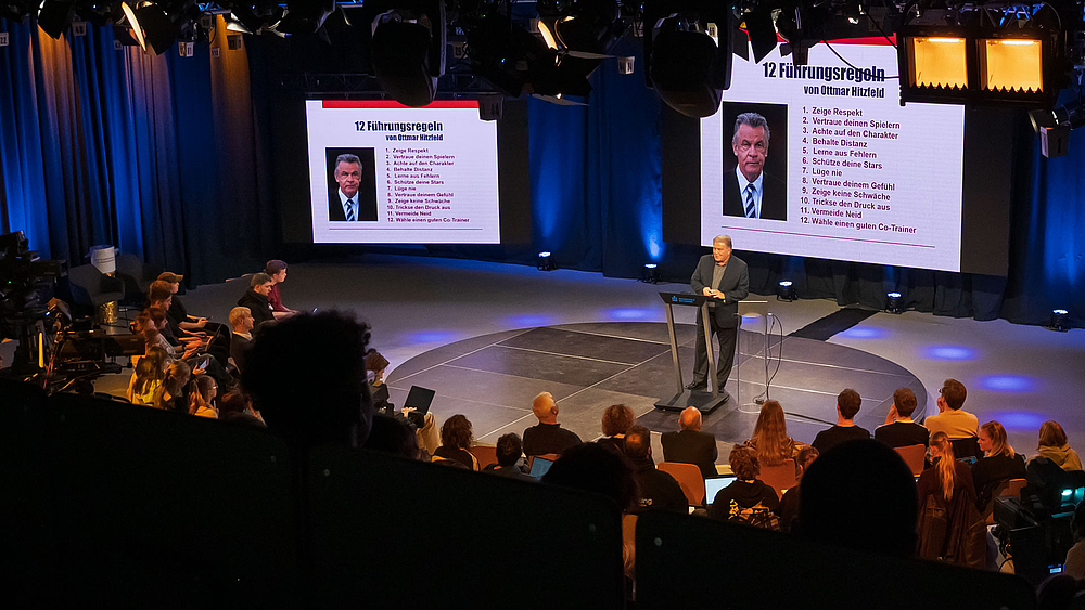 Gelungener Transfer: 12 Regeln für 11 Freunde und ihre Trainer. Blick von oben ins Studio auf den Referenten und die Screens im Hintergrund, auf den die 12 Führungsregeln von Ottmar Hitzfeld aufgelistet sind.