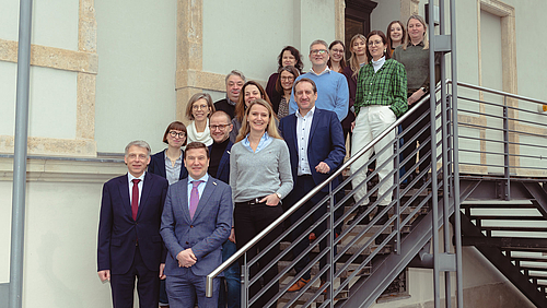 Auf den Stufen zu guter Zusammenarbeit: Die Rektoren der Hochschule Mittweida, Volker Tolkmitt, und der Universität Hasselt, Bernard Vanheusden, (unten v.l.) mit Wissenschaftler:innen aus Mittelsachsen und der belgischen Provinz Limburg. Auf den Stufen einer stählernen Außentreppe zu einem Gebäude stehen 17 lächelnde Personen.