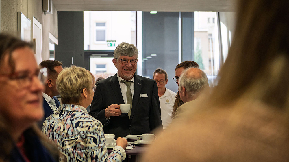 Männliche Person im Anzug lächelnd und mit einer Kaffeetasse in der Hand in einer Gruppe von weiteren Personen an Stehtischen.