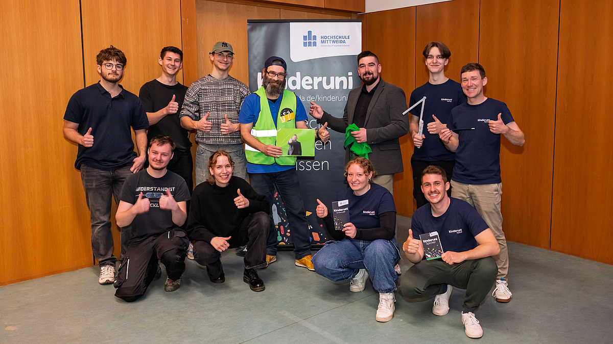 11 lachende Personen mit Daumen hoch vor einem Rollup der Kinderuni.