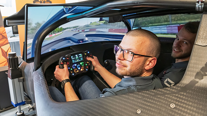 Zwei junge Männer im Cockpit eines Rennwagens, das Teil einer Simulationsplattform ist, in der das Umfeld Rennstrecke auf großen Monitoren dargestellt ist. Der linke Mann fasst mit beiden Händen das Lenkrad. Beide schauen nach hinten in Richtung Kamera. 