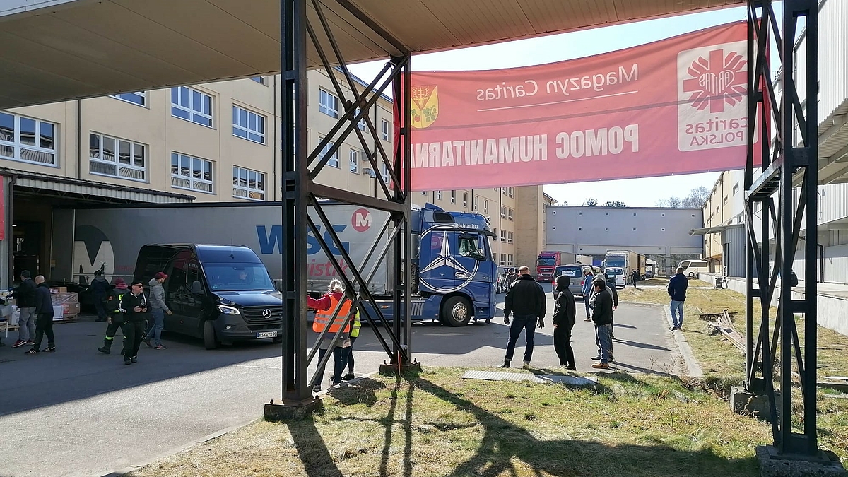 Das Foto zeigt den Innhof eines Gebäudekomplexes. Ein Transparent mit polnischer Beshriftung und dem Caritas-Logo ist von hinten zu sehen. Der LKW aus Mittweida steht an einer Laderampe. Daneben steht ein kleinerer Transporter. Einige Menschen und weitere LKW befinden sich auf dem Gelände.