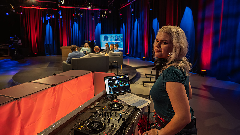 Junge Frau am DJ-Pult im TV-Studio. Im Hintergrund die Personen auf dem Podest.