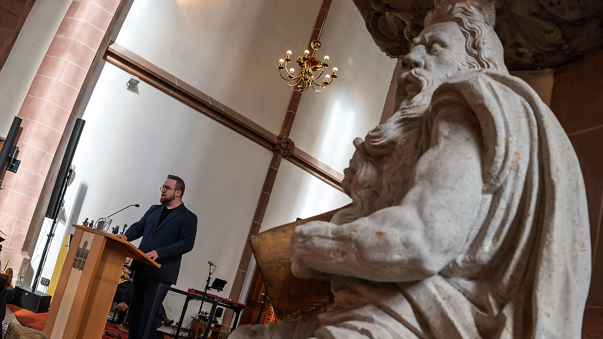 Blick auf den Redner im Hintergrund an einer Mose dartsellenden Sandsteinfigur im Vordergrund vorbei. Redner und Figur verbindet, dass sie einen Vollbart tragen