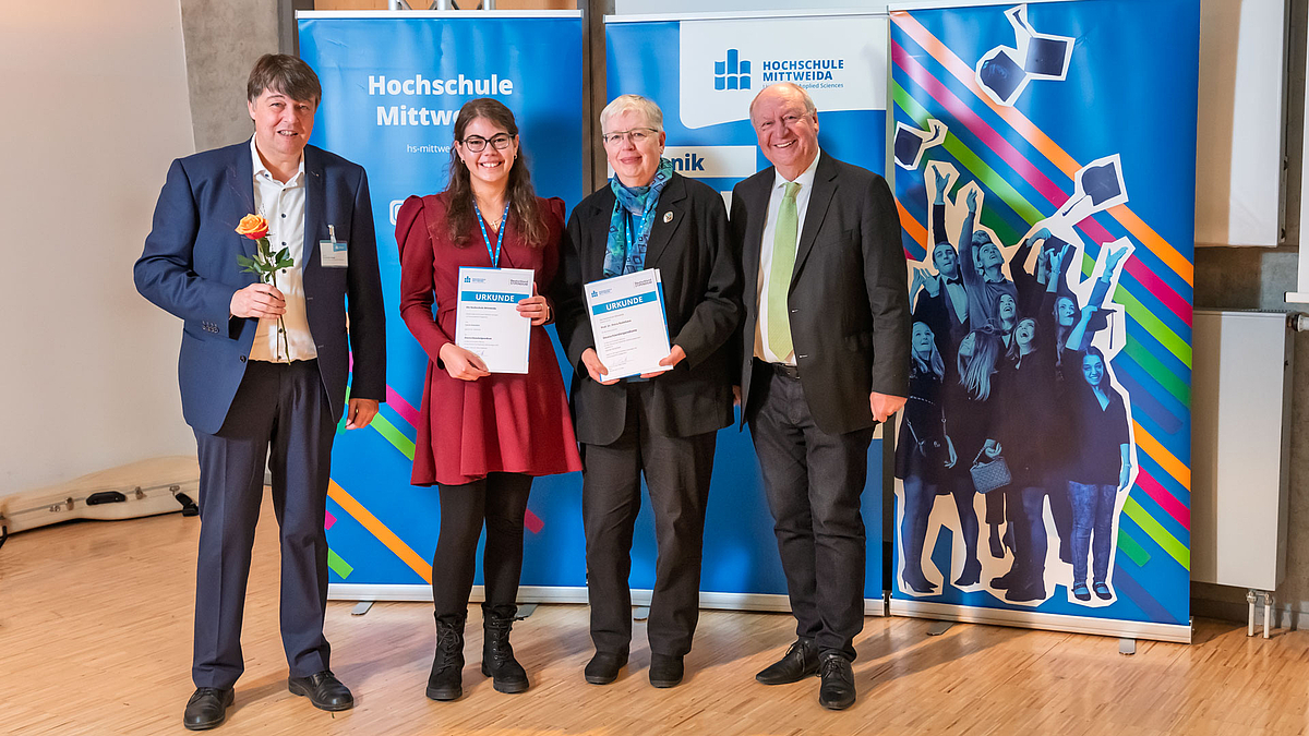 Fördert als Privatperson: Prof. em. Dr. Petra Radehaus, hier mit „ihrer“ Stipendiatin Leonie Kotschick. Die Kombination passt, denn beide sind in der Biotechnologie zuhause. Zwischen zwei lächelnden männlichen Personen, von denen die linke eine orangefarbene Rose hält, stehen zwei lächelnde weibliche Personen, die jeweils eine Urkunde vor sich in den Händen halten. Sie stehen vor drei blauen Rollups mit Logos, Schriftzügen der Hochschule Mittweida und einem typischen Absolventen-Hut-Wurf-Bildmotiv. Der Inhalt der Urkunden ist nicht lesbar.